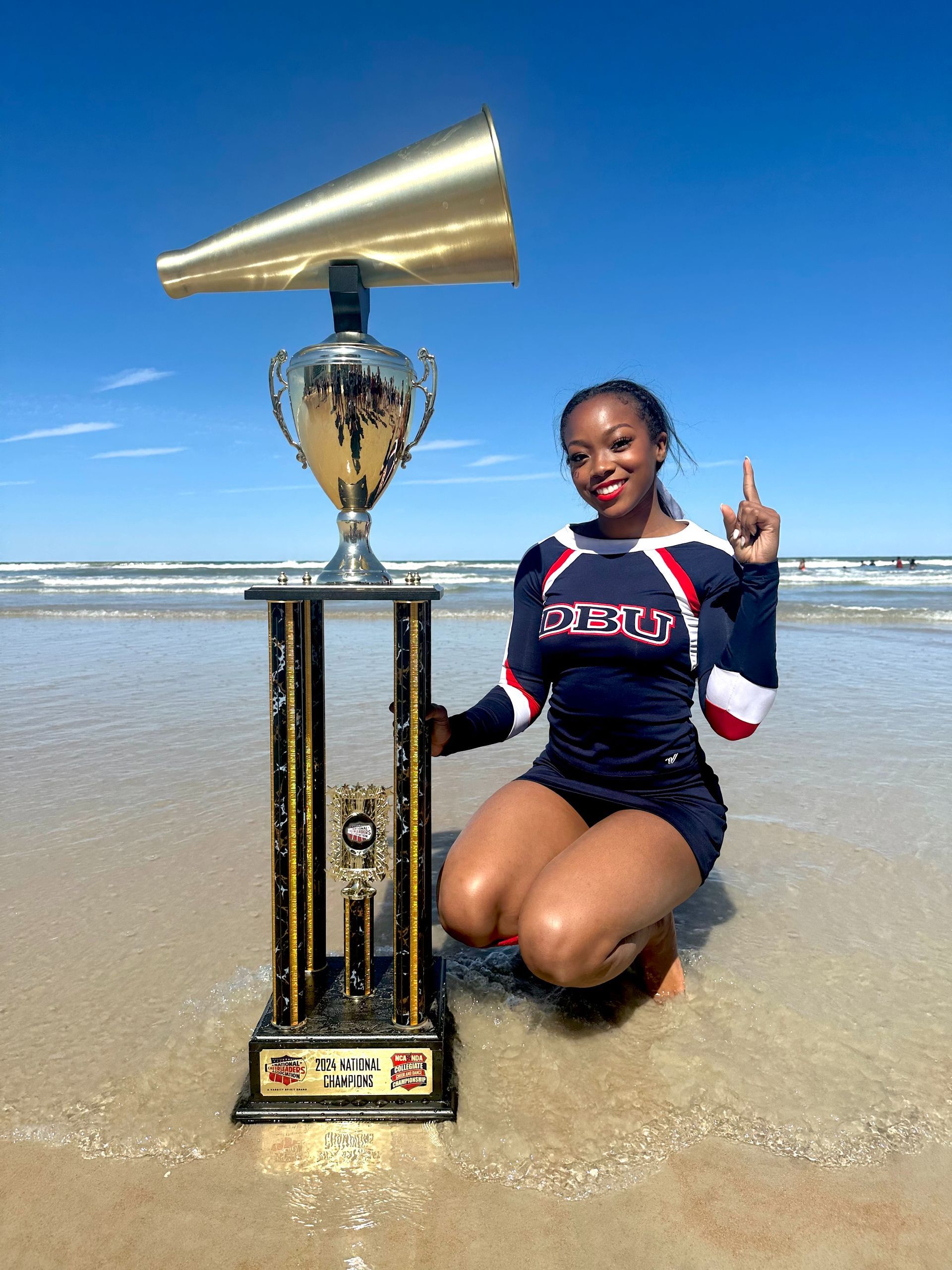 A cheerleader in a dark uniform kneels on a sunny beach next to a large trophy topped with a gold megaphone.