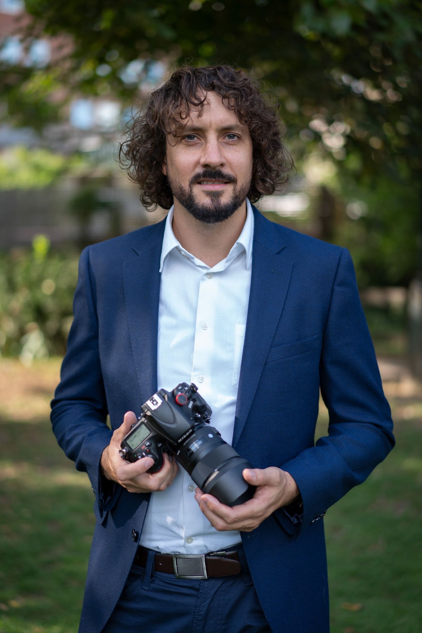 Edward with curly hair in a blue suit, holding a camera, outdoors.
