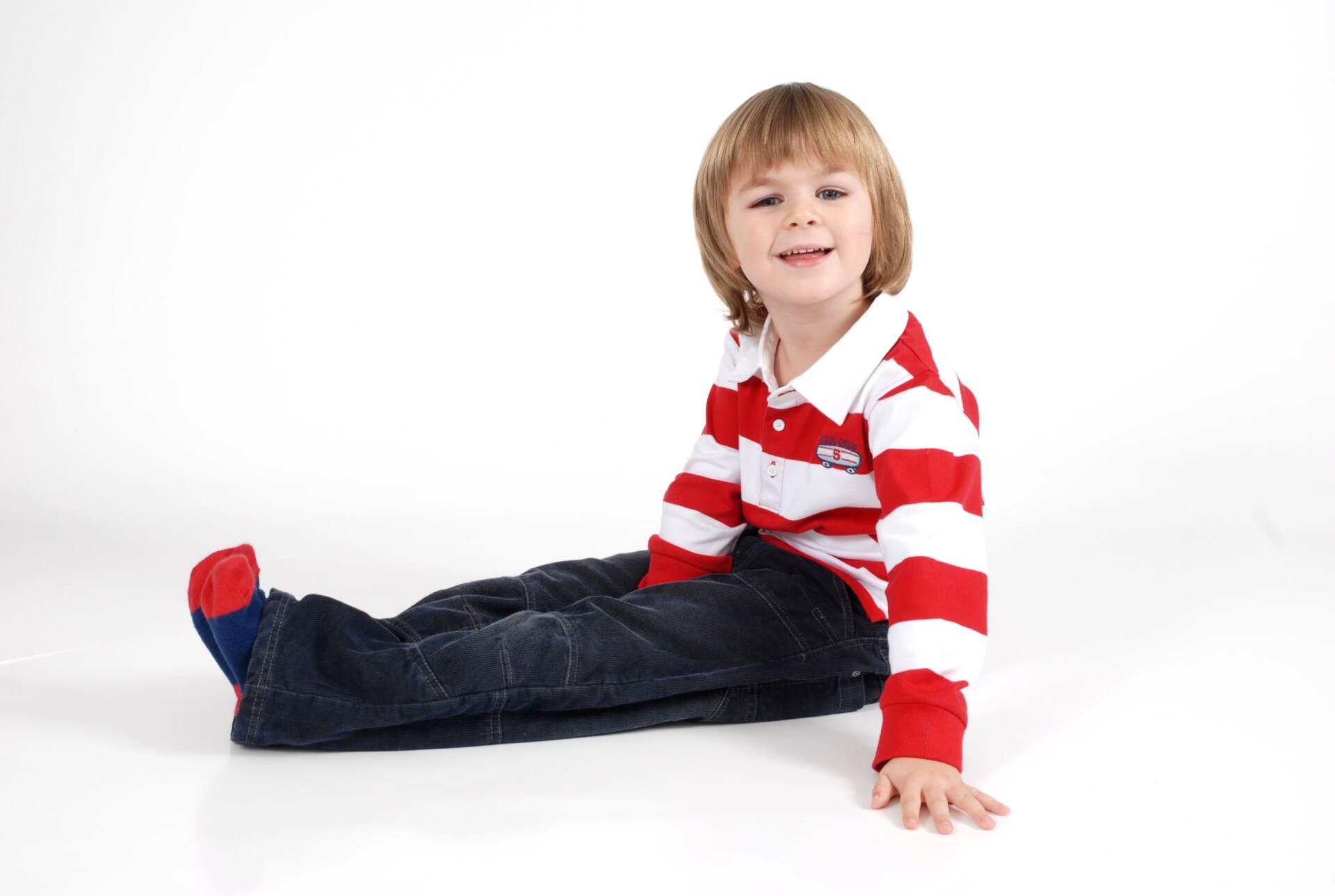 Young boy with blonde hair wearing red/white striped shirt and jeans sits on a white surface.