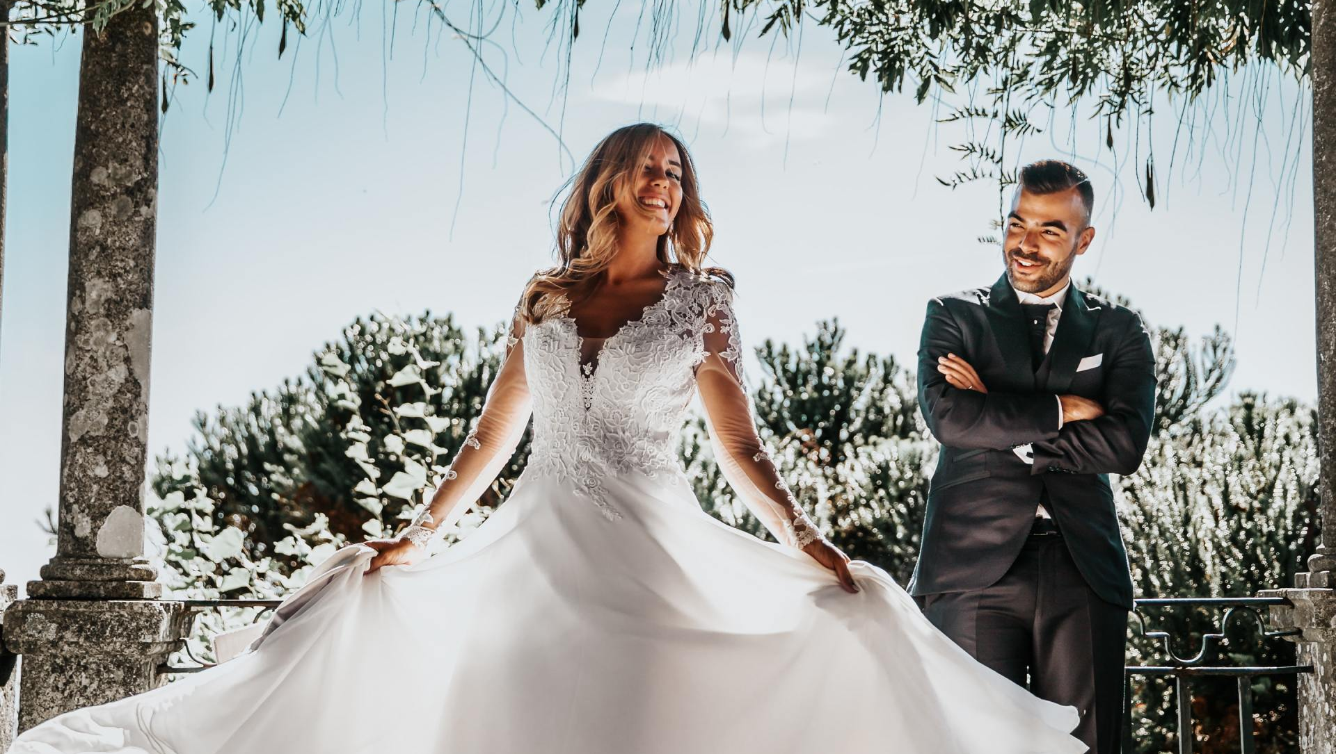 Bride in wedding dress twirls outdoors, groom watches with folded arms. Sunny, joyful.