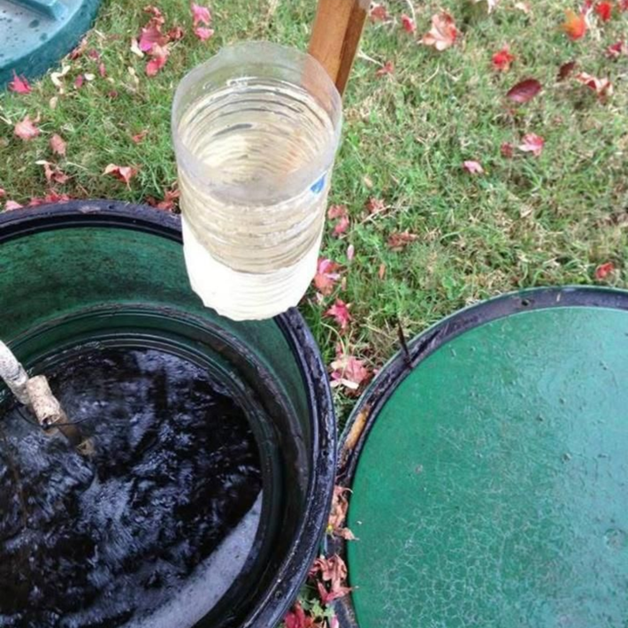 A plastic bottle with liquid hangs over an open septic tank. Green lids on the ground, grass in background.