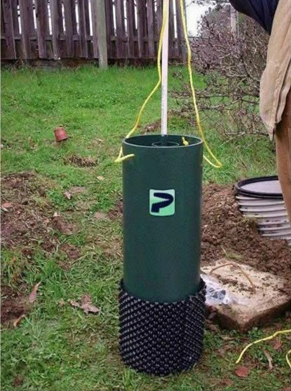 Green well casing with a black base, yellow wires, and a white pole in a grassy yard.