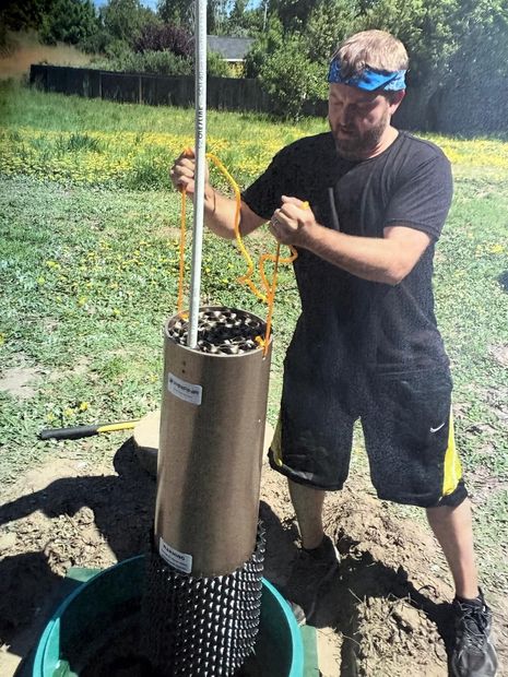 Man lowering a cylindrical object into a well. He’s wearing a black shirt and shorts, and is outdoors.