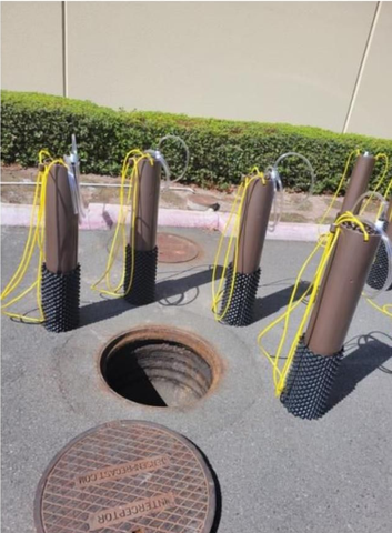Five brown cylinders with yellow wires are placed around an open manhole in asphalt.