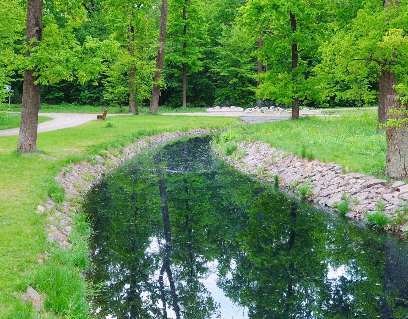 A narrow, reflective stream winds through a green park, lined with trees and grass.