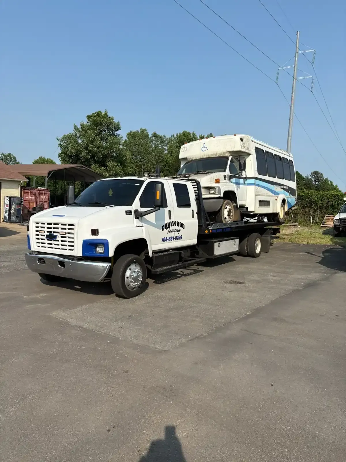 White tow truck carrying a small bus on its flatbed, parked outdoors.