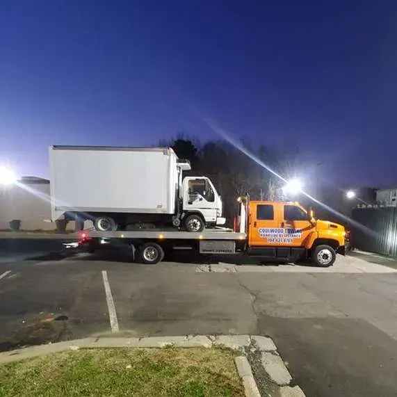 Orange tow truck carrying a white box truck on a flatbed at night.
