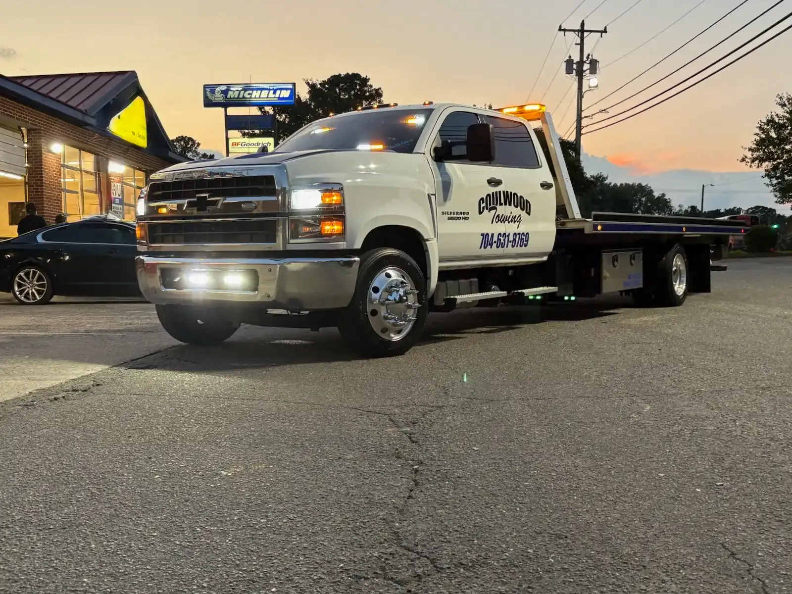 White tow truck with emergency lights on parked in front of a building at dusk.