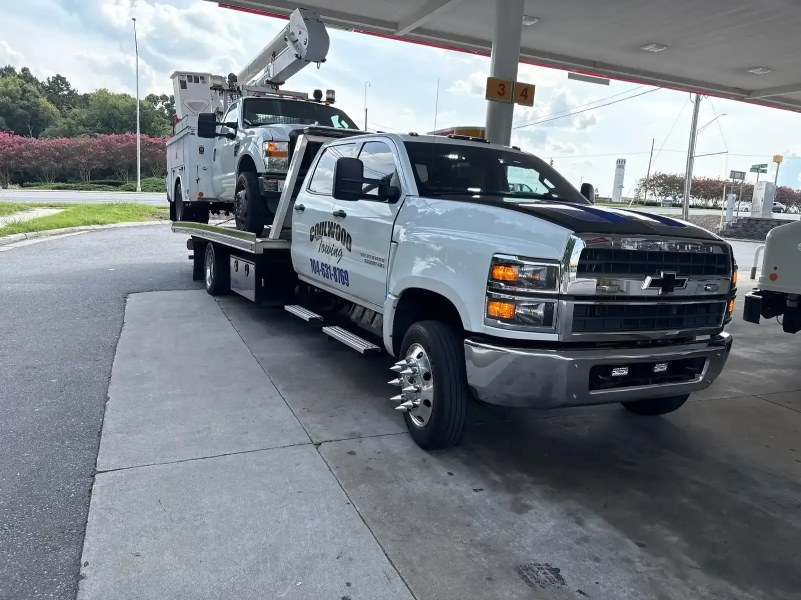 A white tow truck with a smaller utility truck on the bed, parked under a gas station awning.