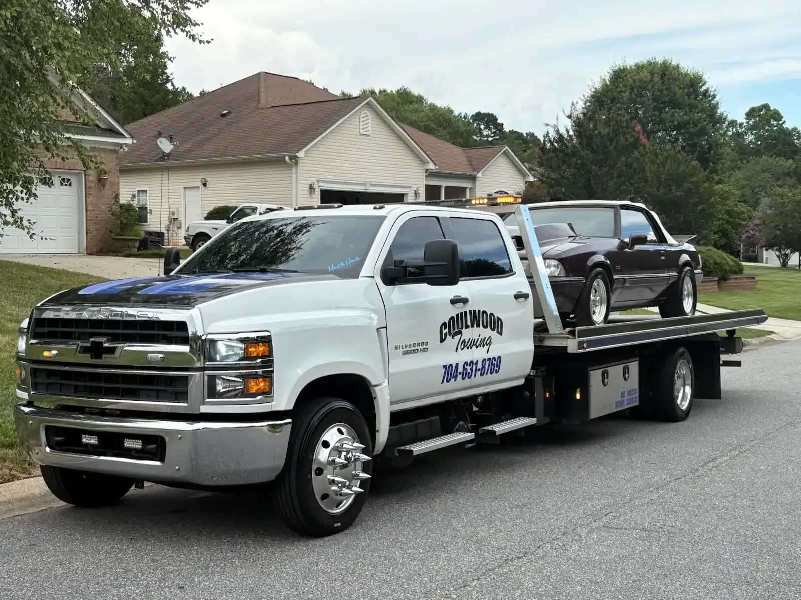 Tow truck with a maroon car on its bed, parked in front of houses on a sunny day. The truck is white with black lettering.