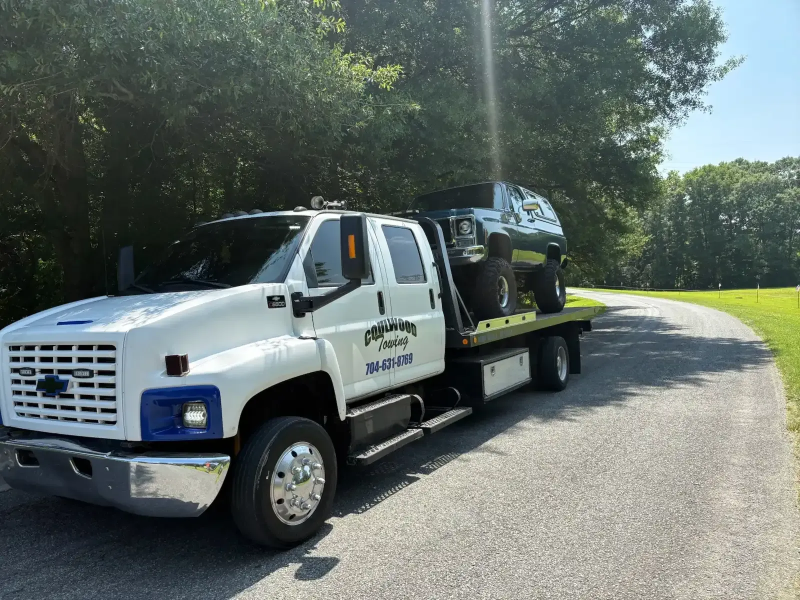 White tow truck carrying a black SUV on a paved road surrounded by trees.