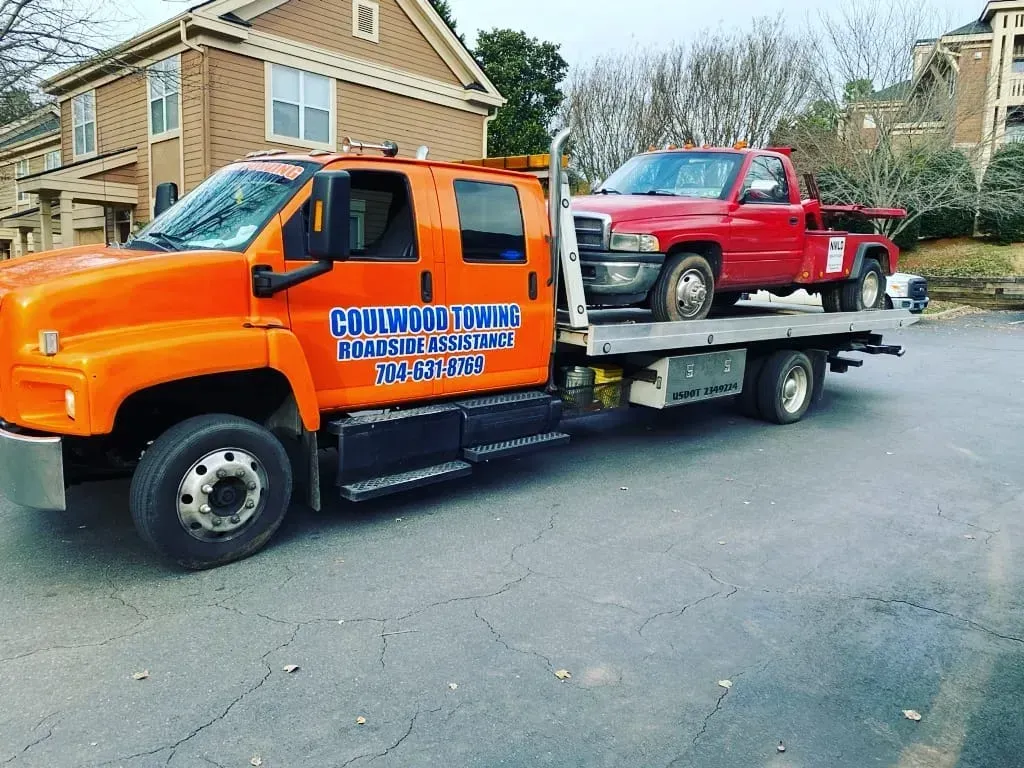Orange tow truck carrying a red pickup truck. Buildings in the background.