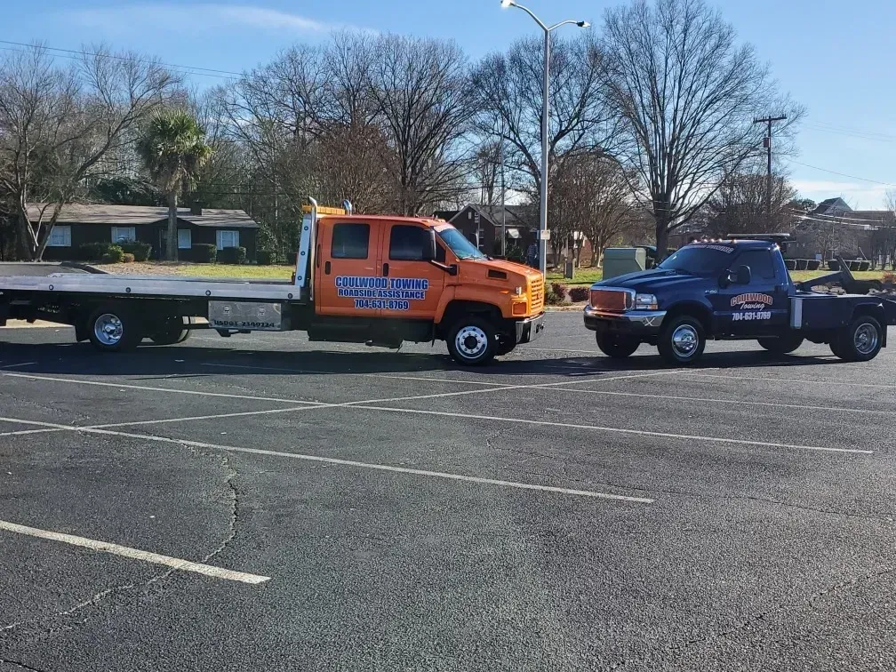 Two tow trucks, one orange and one blue, parked in a lot on a sunny day.