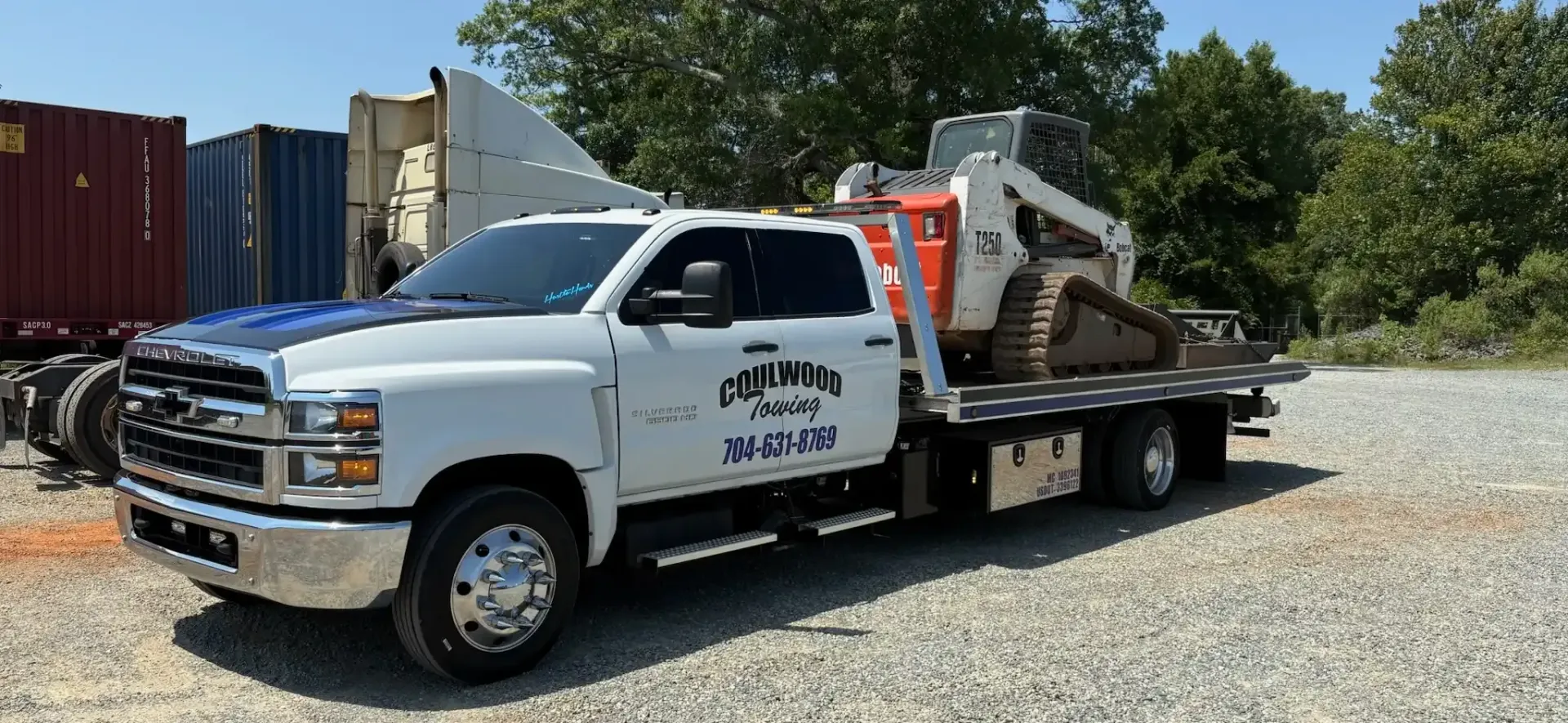 White tow truck with a small excavator on its flatbed. Outdoors, trees in the background.