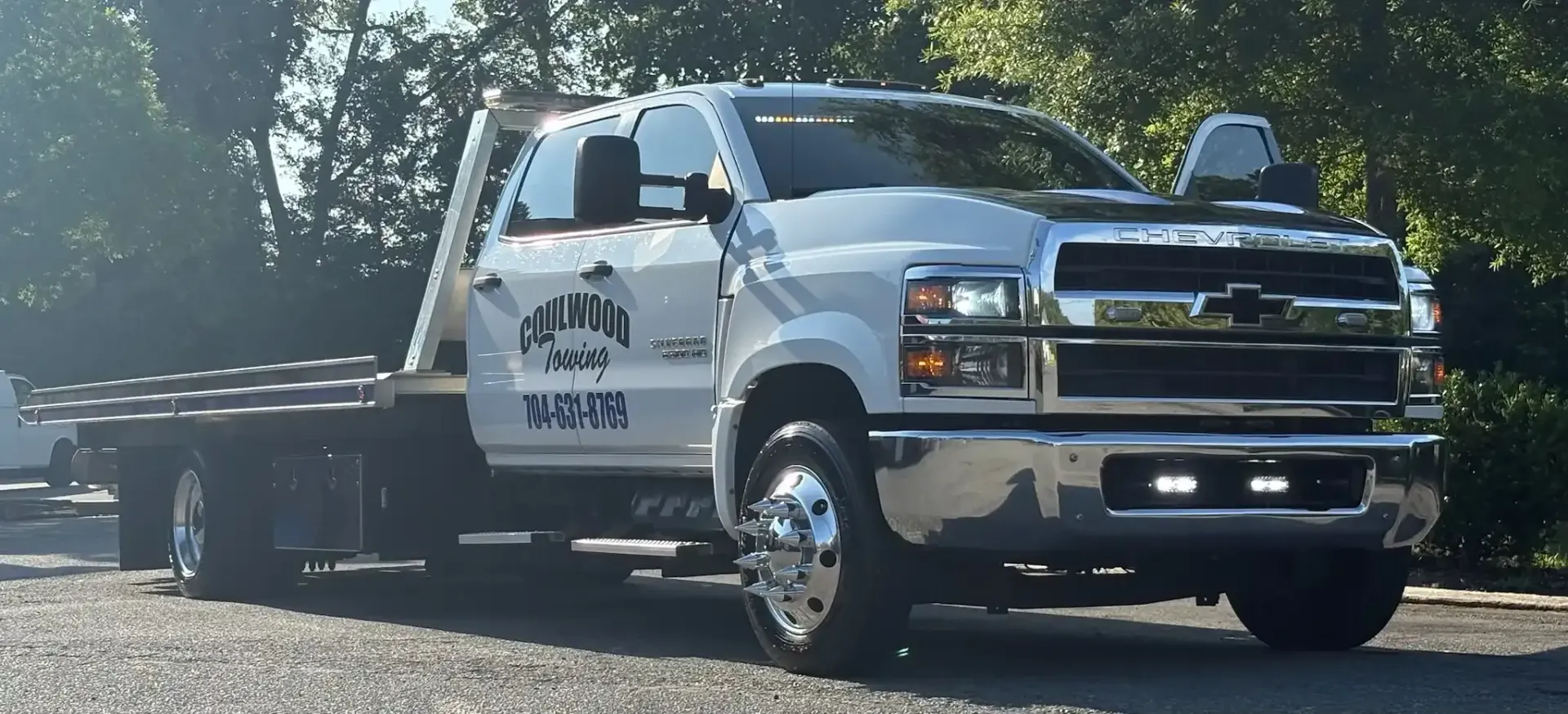White tow truck parked on a gravel surface. The truck has a flatbed. Trees in the background.
