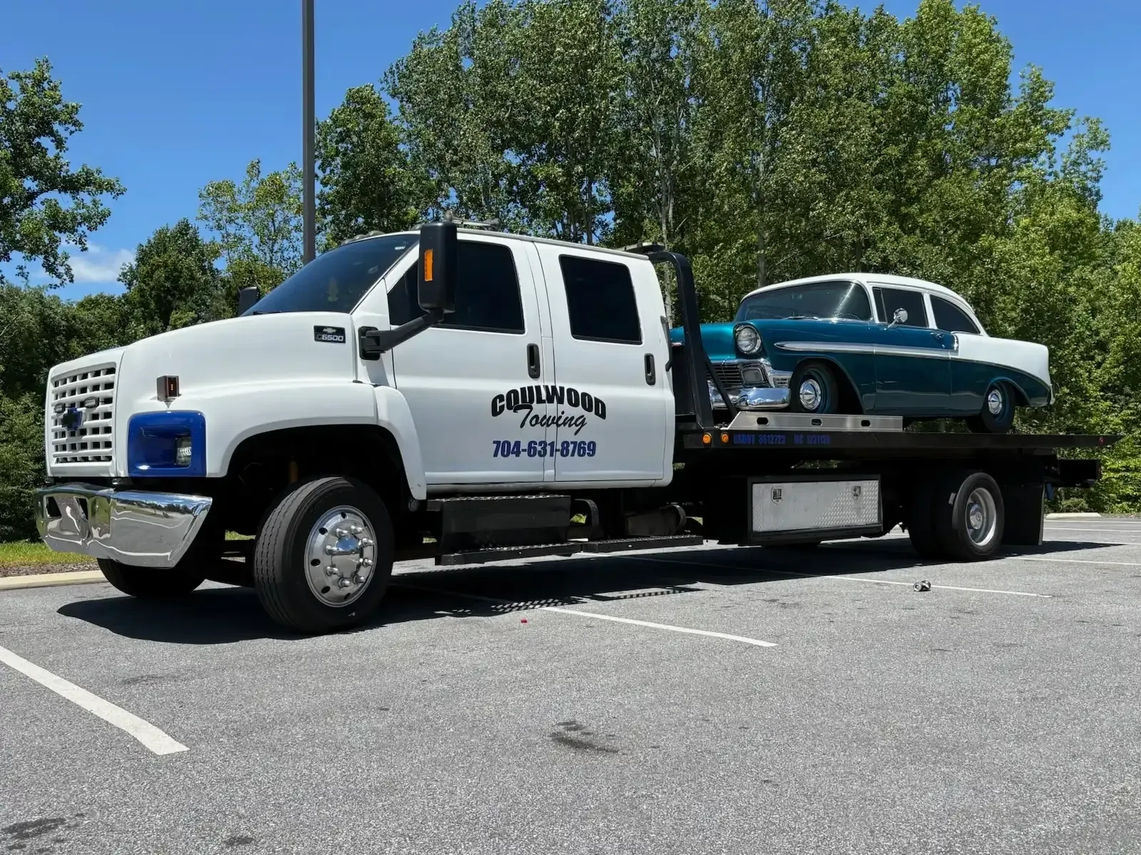 White tow truck hauling a vintage teal and white car on a sunny day.