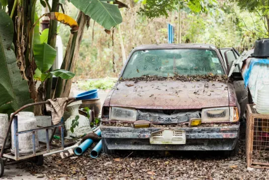 Old, dusty car parked among debris, brown with leaves on the hood.