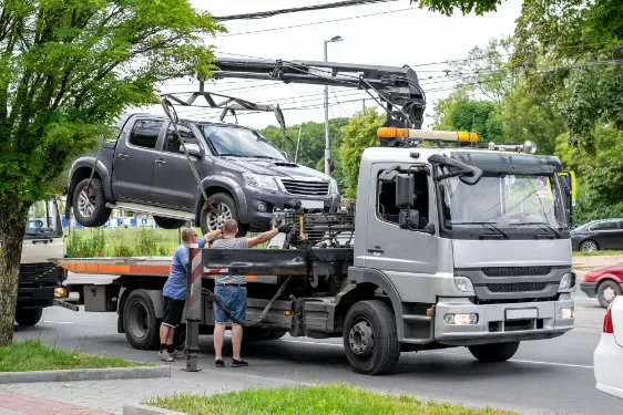 Tow truck lifting a gray pickup truck; two people watch on a street.