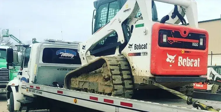 Bobcat track loader, muddy, on a tow truck. White and orange machine, cloudy day.
