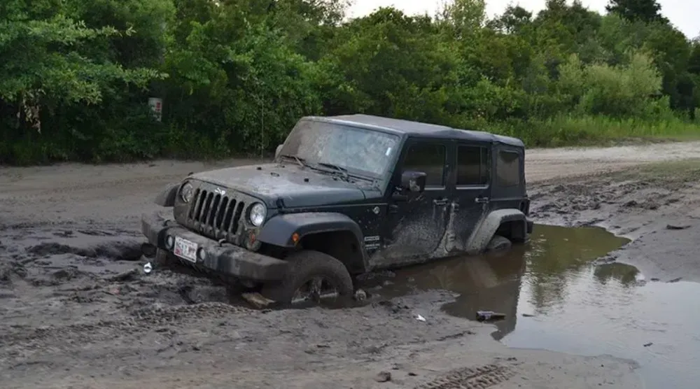 Dark Jeep Wrangler stuck in deep mud on a dirt road, near greenery.