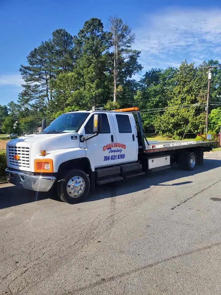 White tow truck parked on a paved road; the truck has a flatbed and the words 