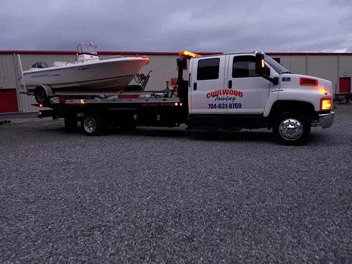 Tow truck transporting a white boat on a trailer, outside a building, dusk.