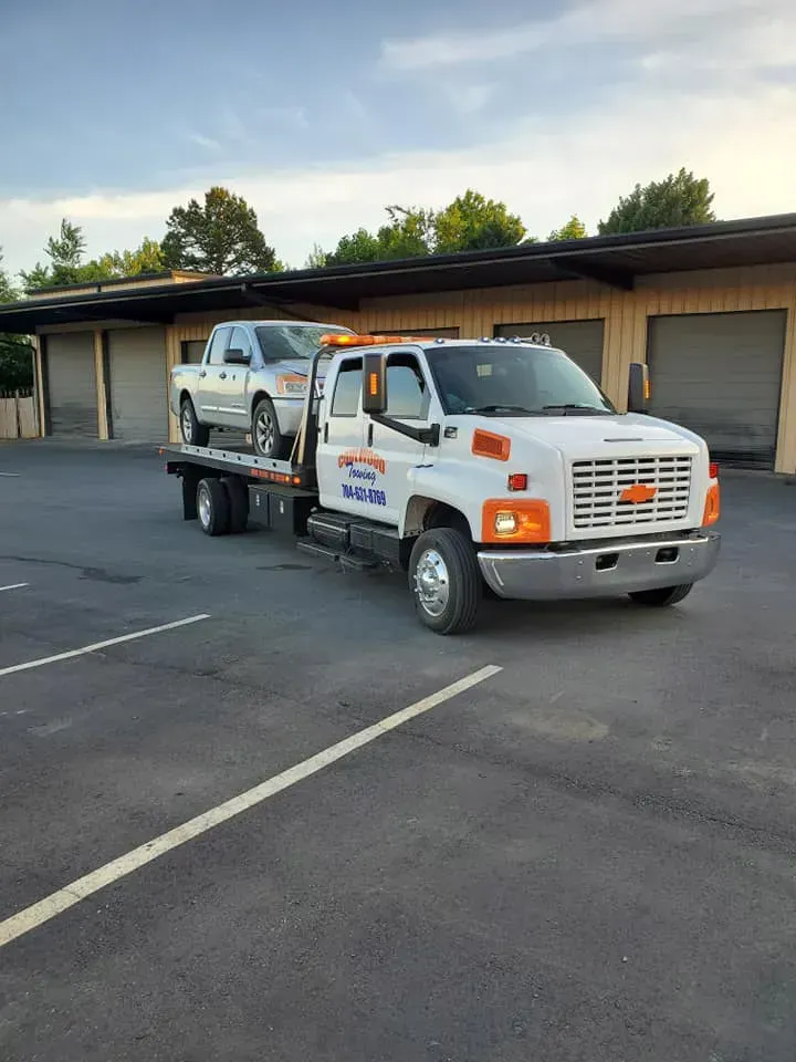 Tow truck carrying a pickup truck in a parking lot. White truck, blue sky, buildings.