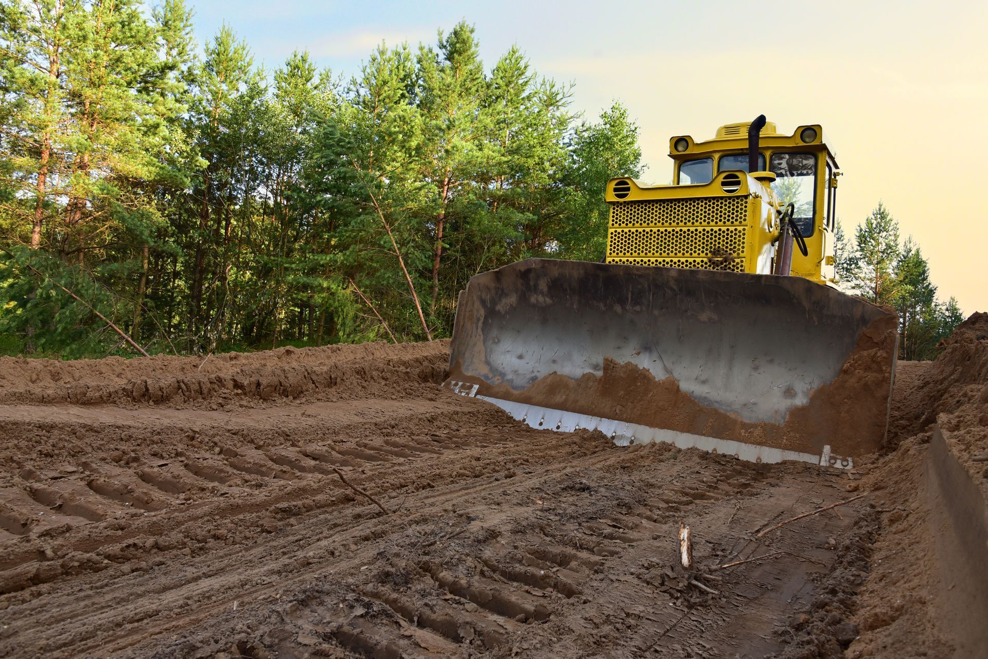 Yellow bulldozer leveling muddy dirt road beside trees under a clear sky