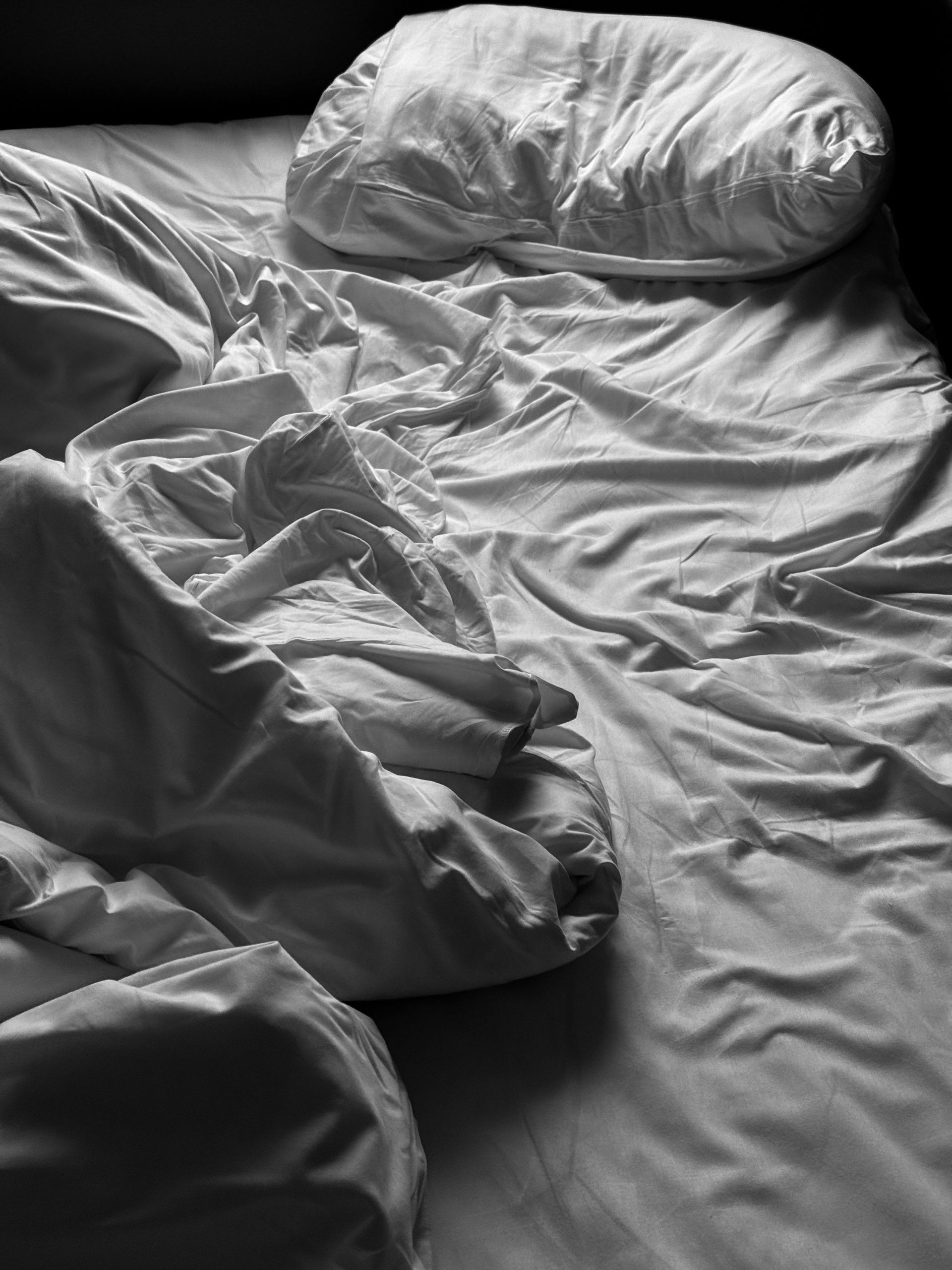 Unmade bed with wrinkled gray sheets and a pillow in soft window light