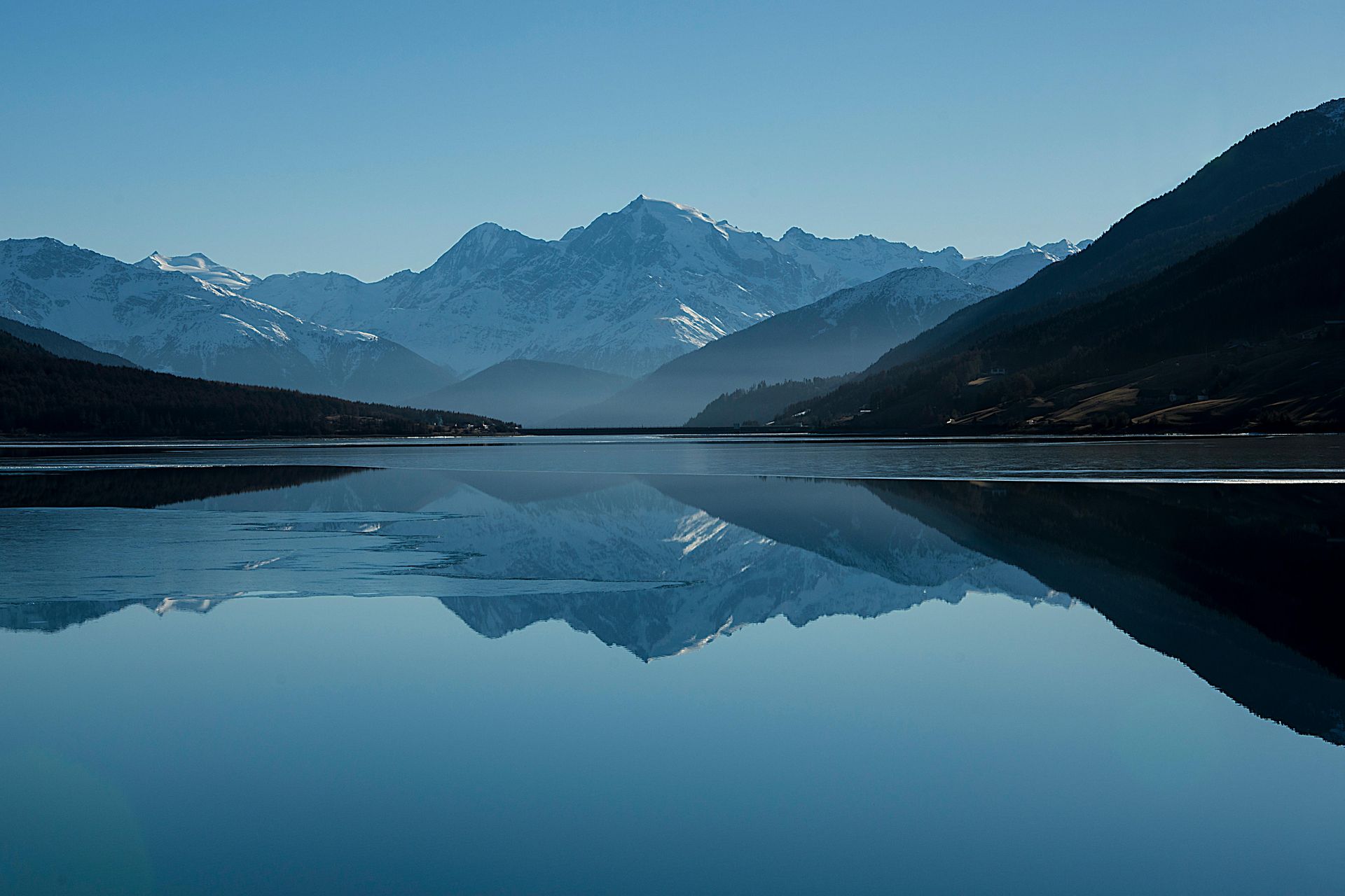 Mountains reflected in a calm lake under a clear, blue sky. Snow on peaks.