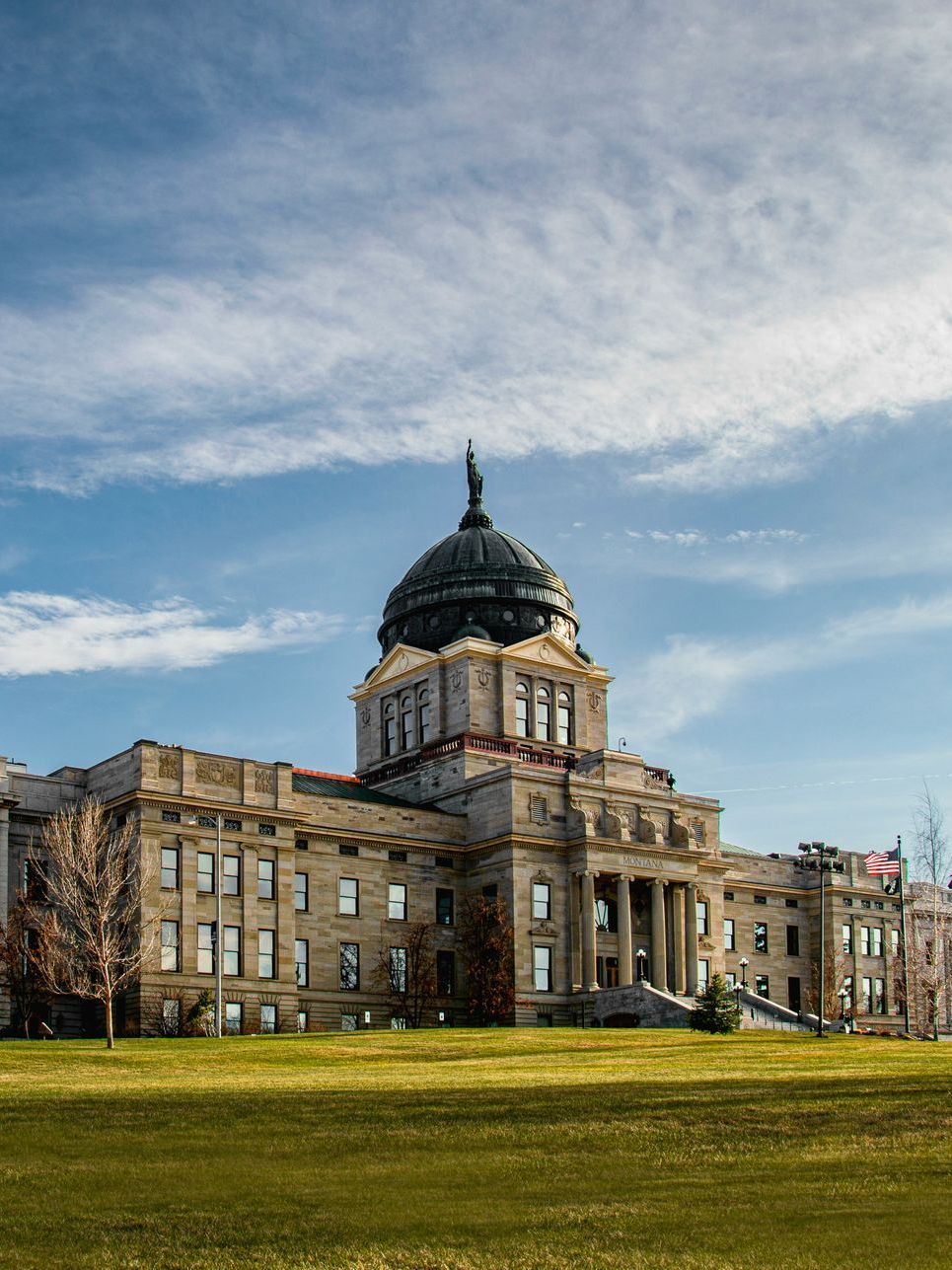 Montana State Capitol building against a blue sky, viewed from a grassy lawn. A large dome crowns the stone structure.