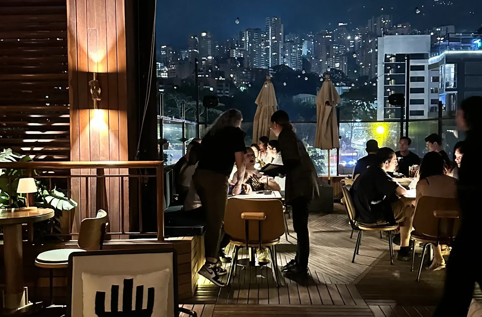 Restaurant patio at night; people dining, cityscape in background, server attending a table.