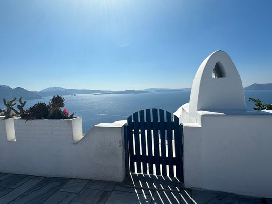 A blue wooden gate sits in a white stone wall overlooking the sparkling blue sea on a sunny day in Santorini, Greece.