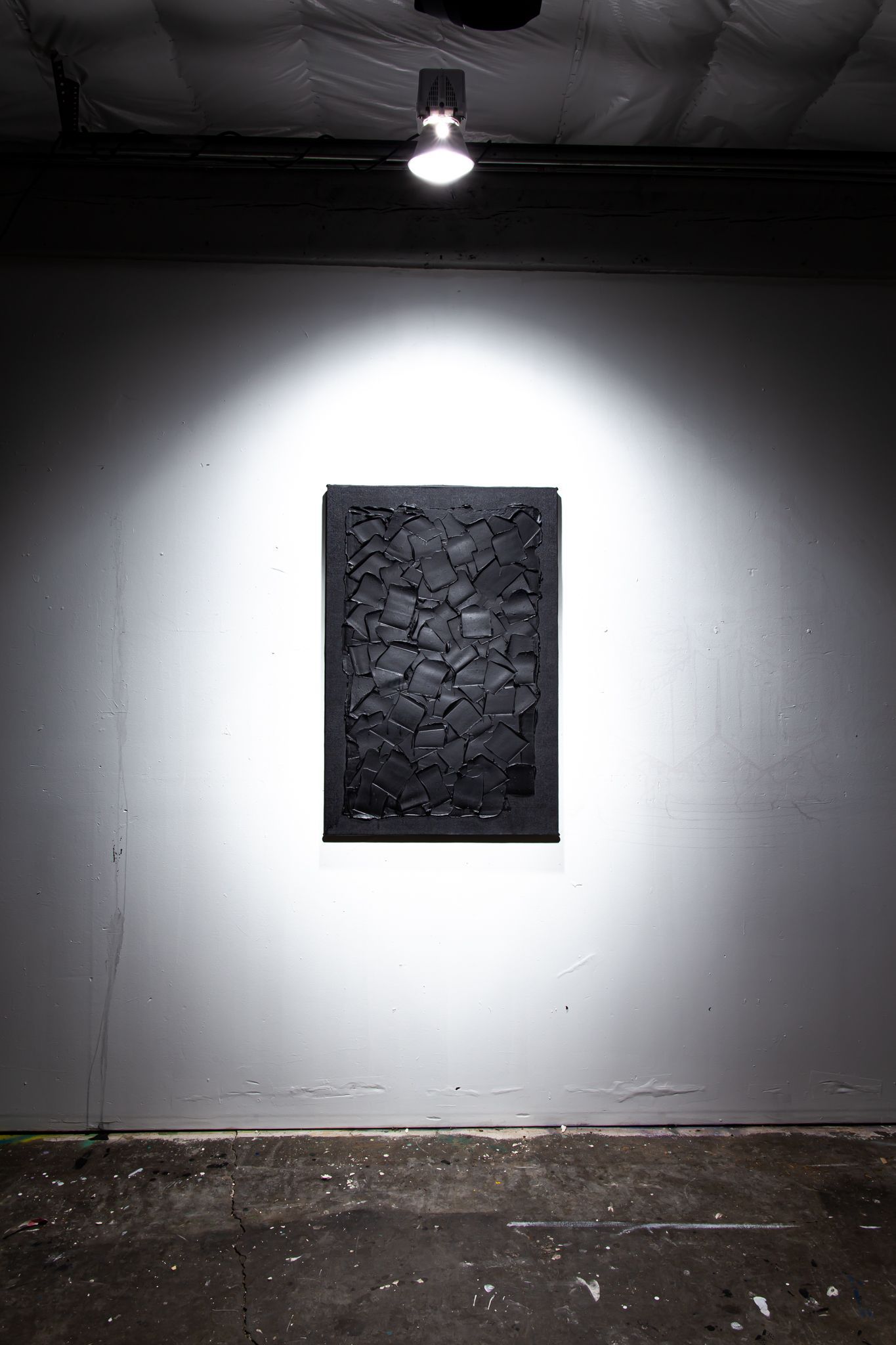 Black abstract artwork on a gray wall, lit by a spotlight in a dimly lit space; art by Matt Remsbecher. 