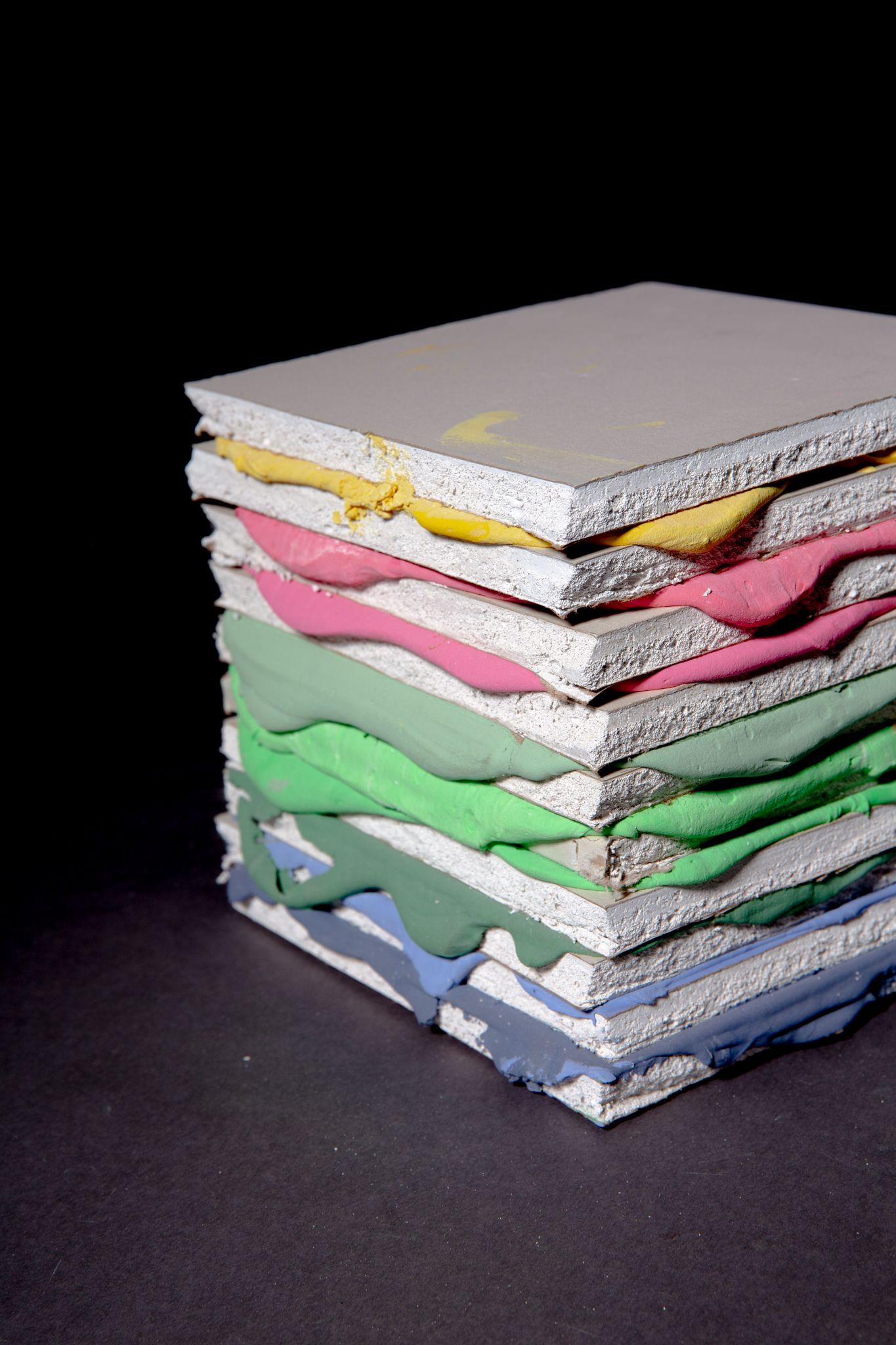 Stack of gray panels, each separated by a layer of colorful material: yellow, pink, green, and blue; by Matt Remsbecher. 