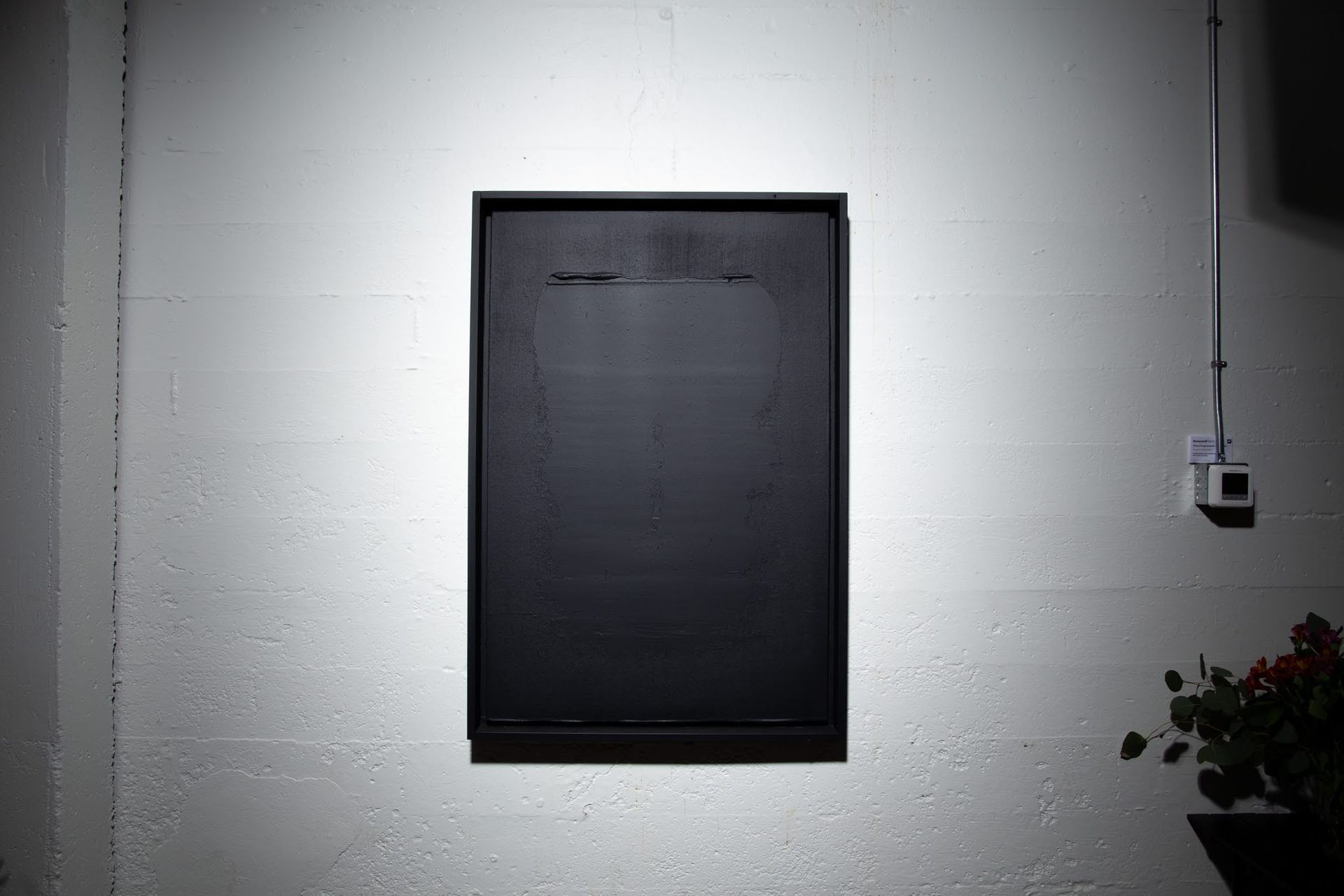 Black rectangular artwork on a white wall; a light switch and plant are in the frame; art by Matt Remsbecher. 