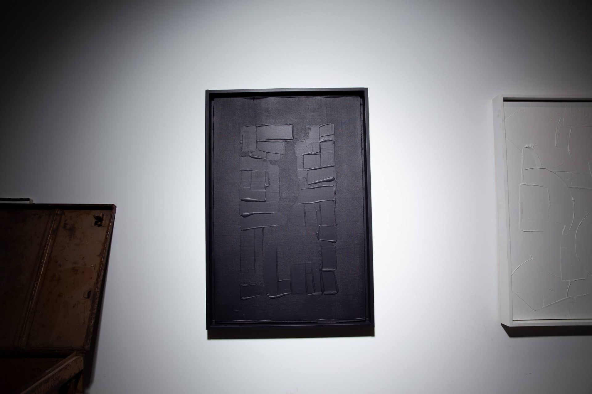 Black abstract artwork in a black frame, on a white wall, under a spotlight; art by Matt Remsbecher. 