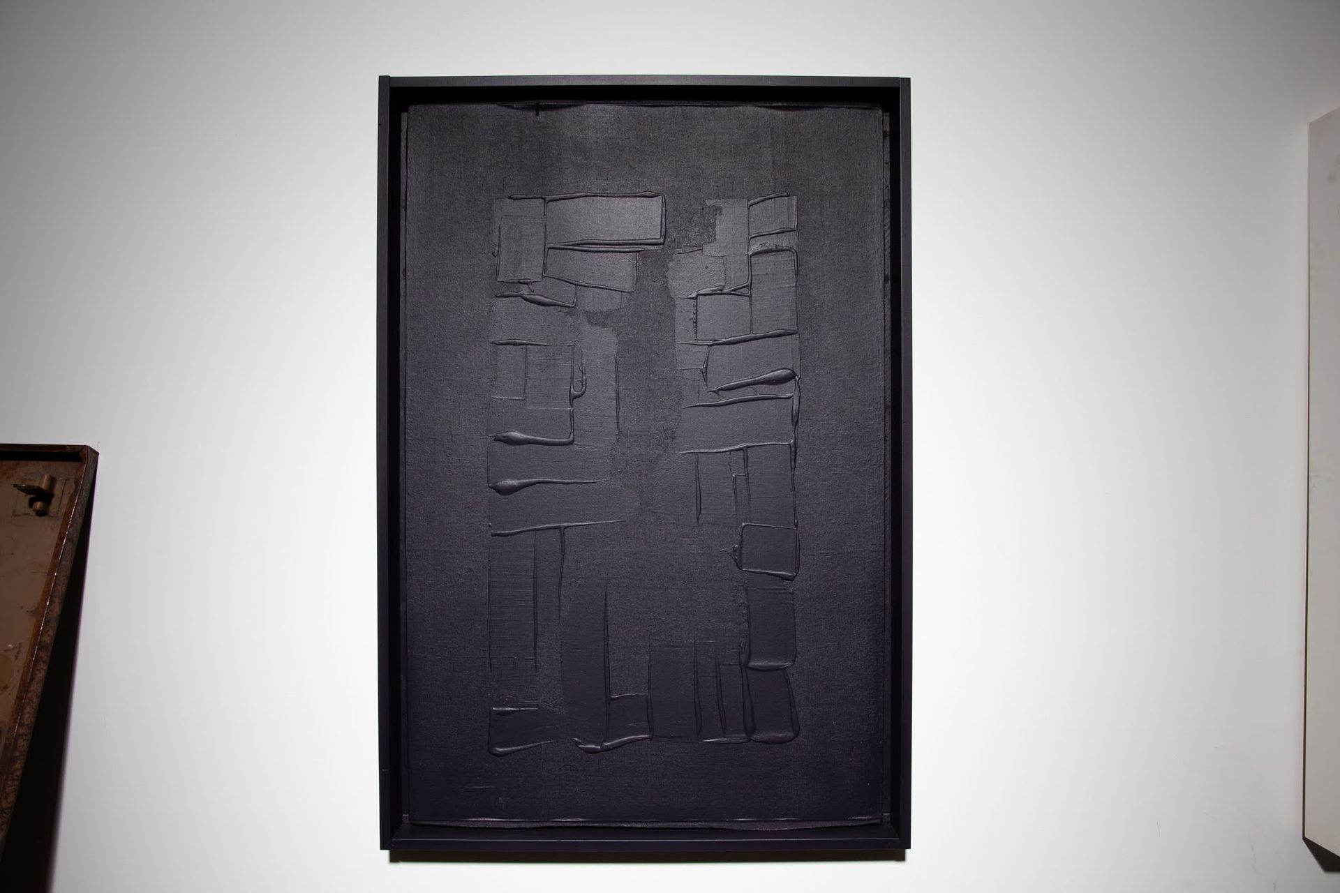 Abstract black rectangular artwork with textured surface in a black frame, hanging on a white wall; art by Matt Remsbecher. 