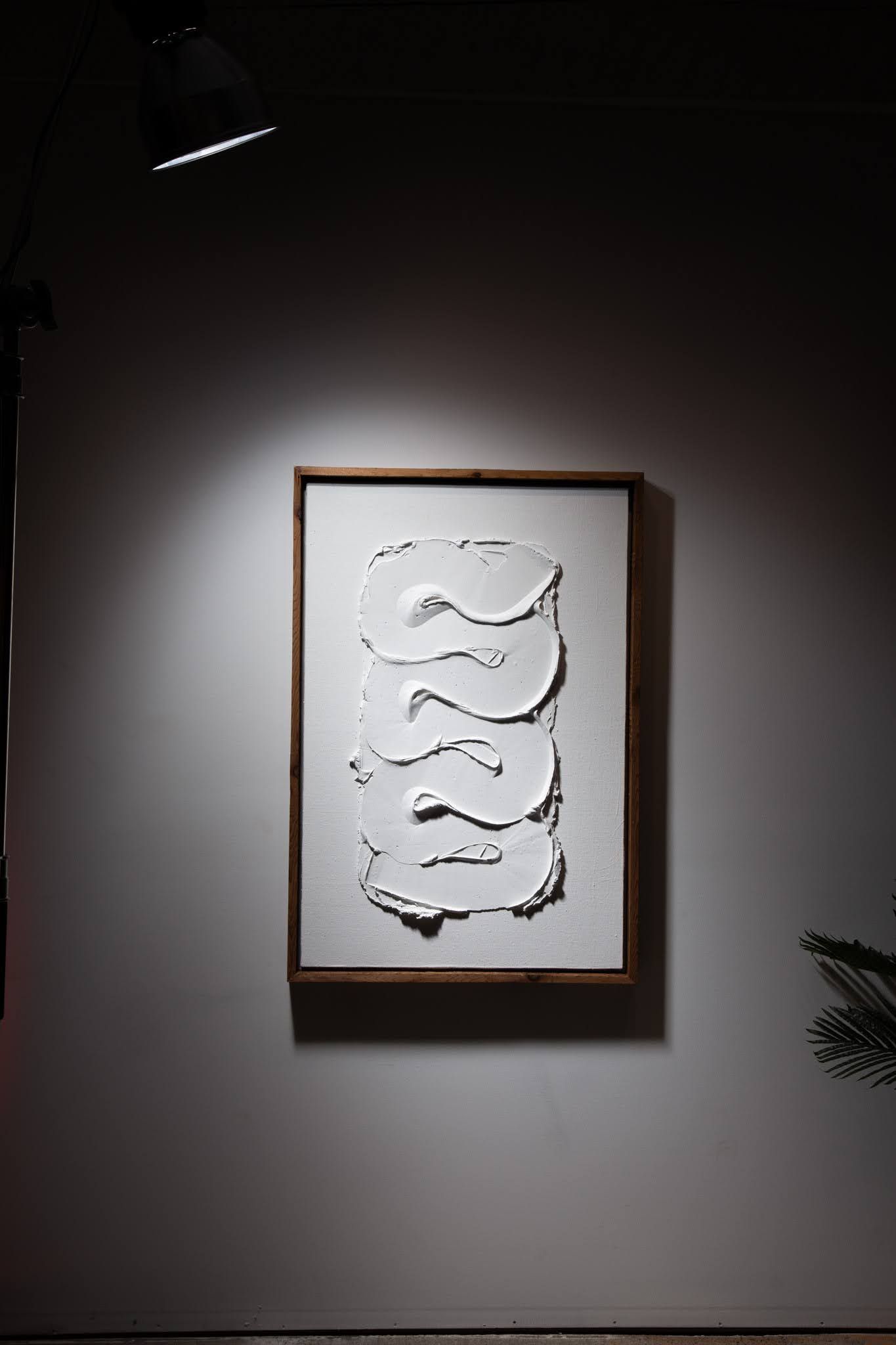 White relief artwork in wooden frame on white wall, lit by spotlight; art by Matt Remsbecher. 