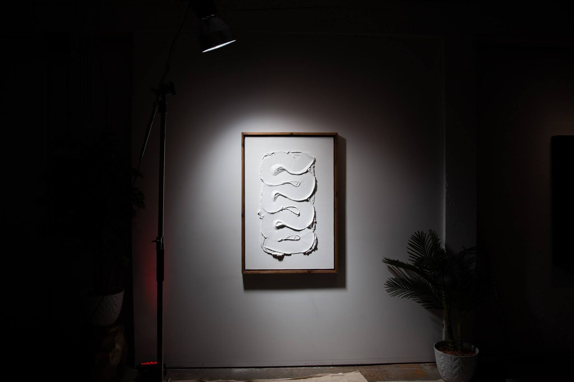 White abstract art in a wooden frame lit by a spotlight on a gray wall; art by Matt Remsbecher. 