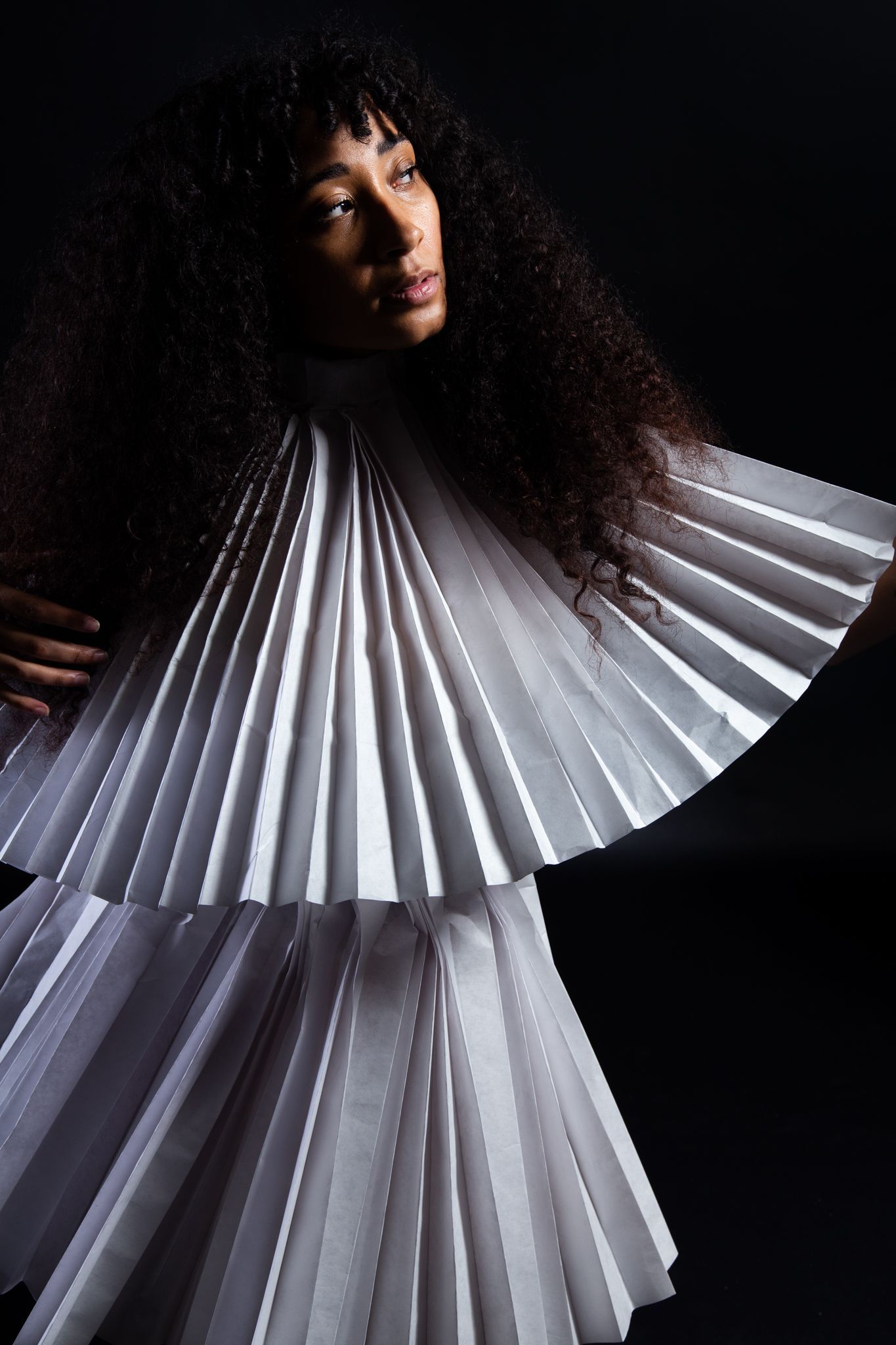 Woman in white pleated garment, looking upward with dark curly hair against a black backdrop; art by Matt Remsbecher. 