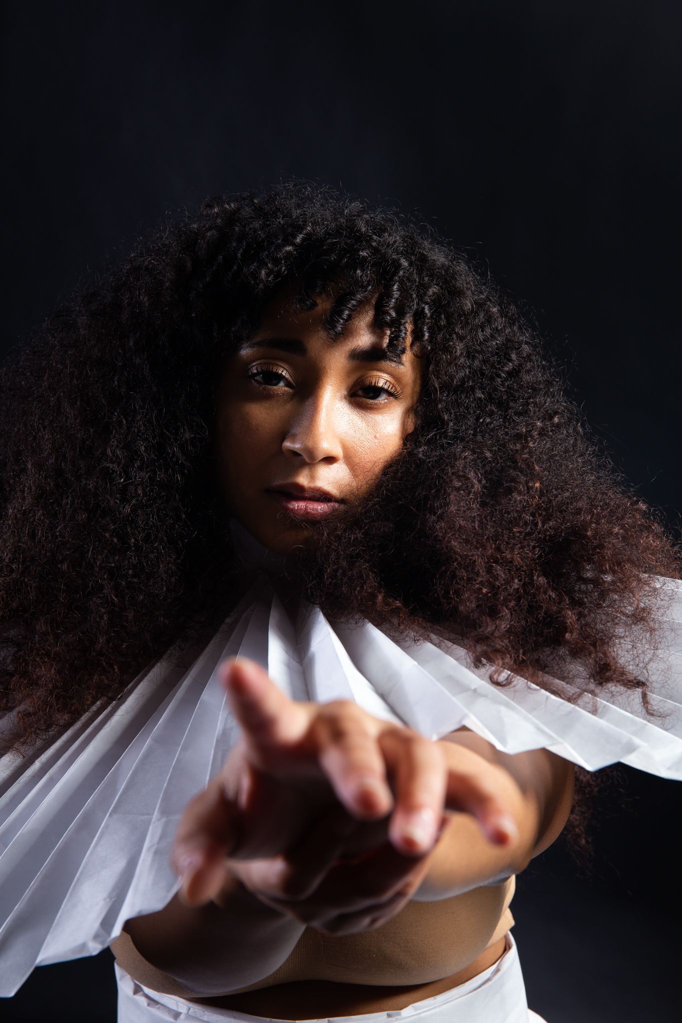 Woman with curly hair, white collar, extending hands towards camera, dark background; art by Matt Remsbecher. 