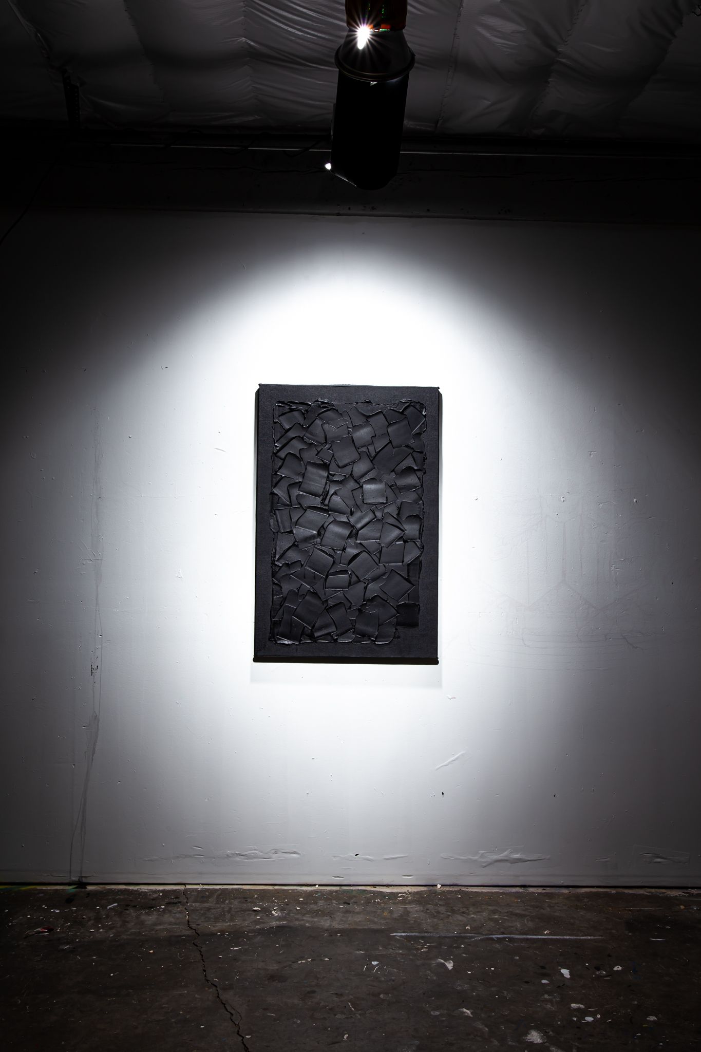 Dark artwork on a wall, illuminated by a spotlight. Textured black surface, in a dimly lit space; art by Matt Remsbecher. 