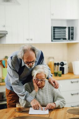 An elderly couple is sitting at a table in a kitchen looking at a piece of paper.