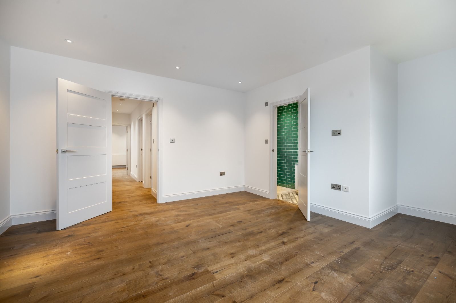 Empty room with wood floors and white walls, two open doors leading to hallways.