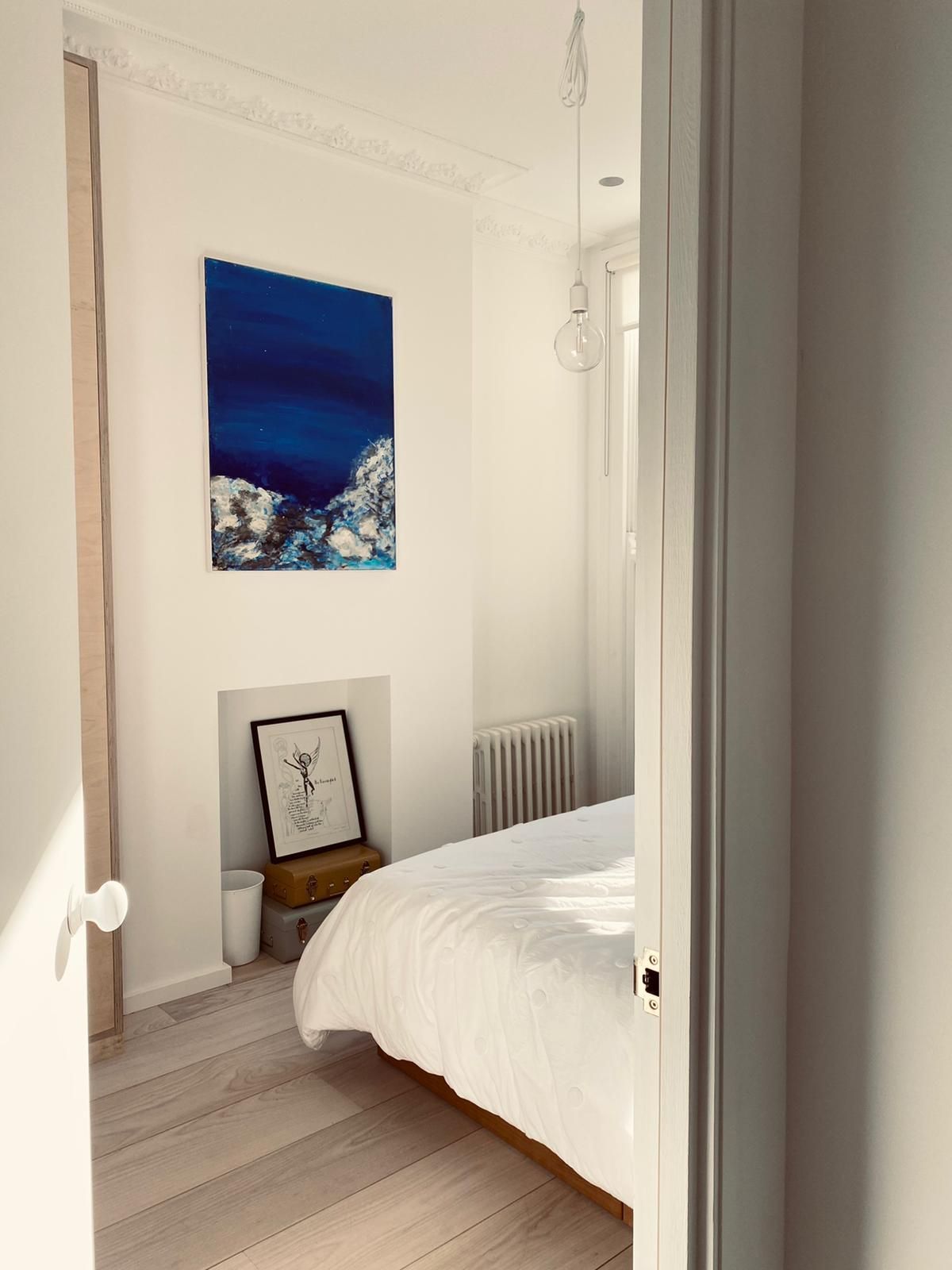 Bedroom with white walls, light wood floor, and a bed with white linens. Blue artwork hangs on the wall.