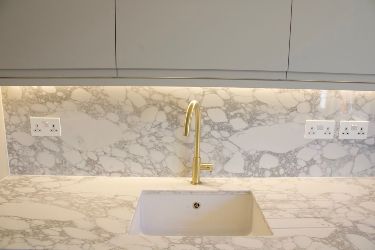 Gold faucet over a white sink in front of marble backsplash and countertop with overhead cabinets and electrical outlets.