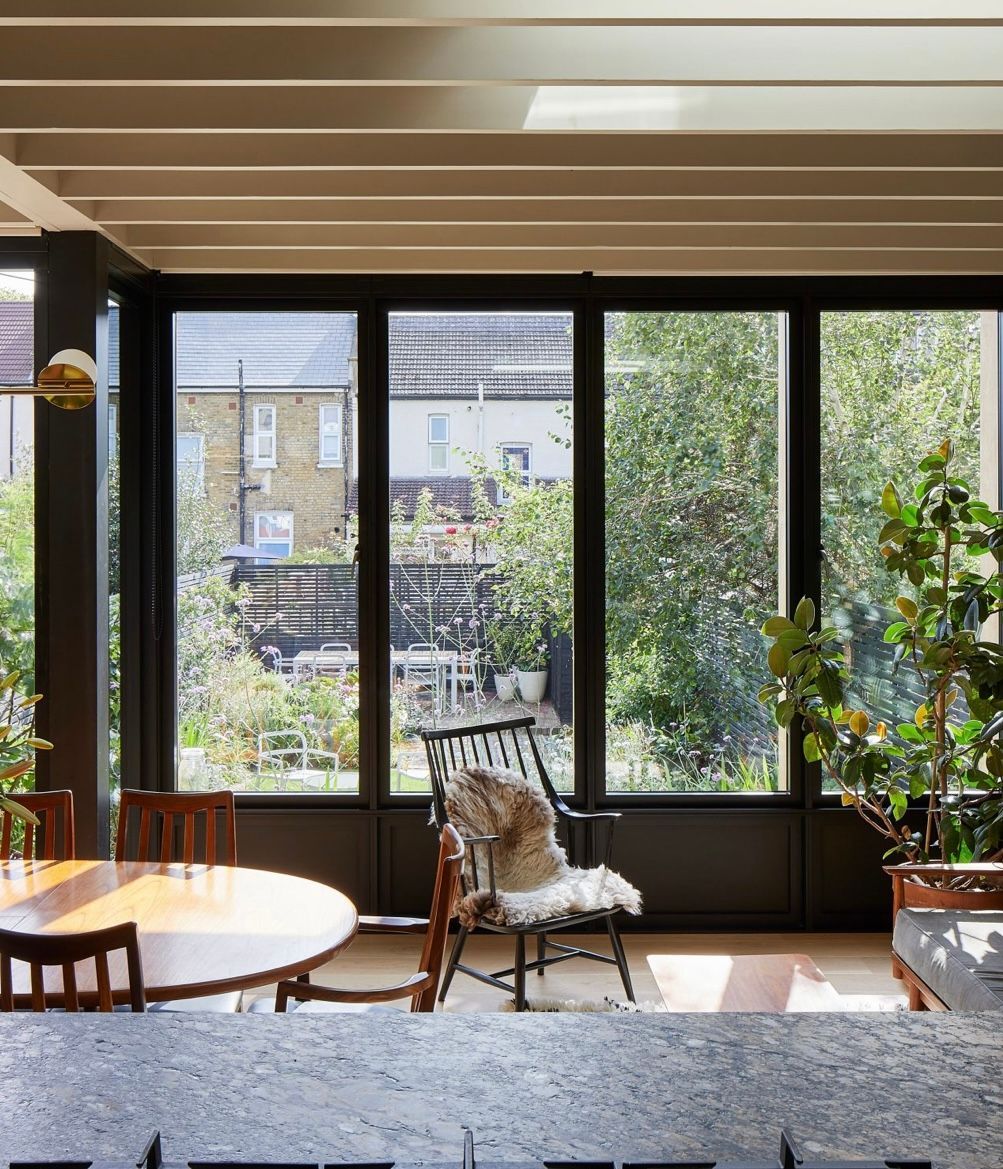 Dining room with large windows overlooking a backyard with a table, chairs, and plants.