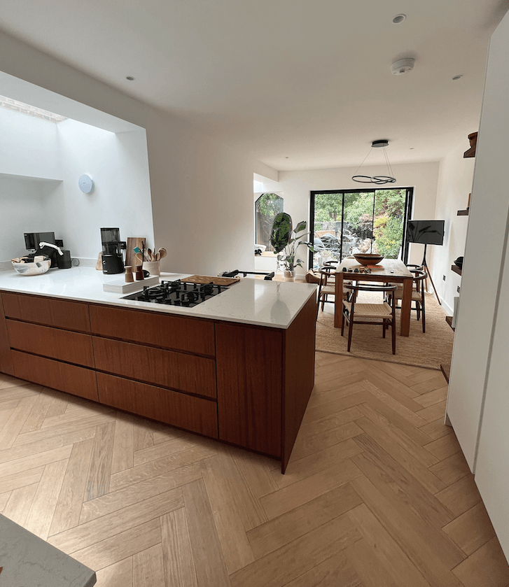 Modern kitchen with wooden island, white countertops, and herringbone floors leads to dining area.
