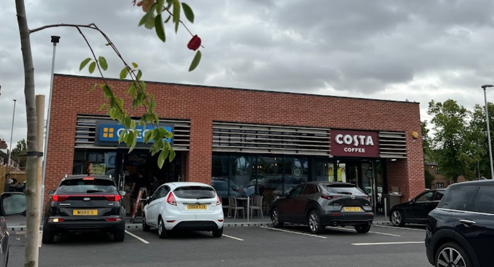 Exterior view of a Costa Coffee shop with parked cars. The building is brick with the Costa Coffee logo visible. Overcast sky.